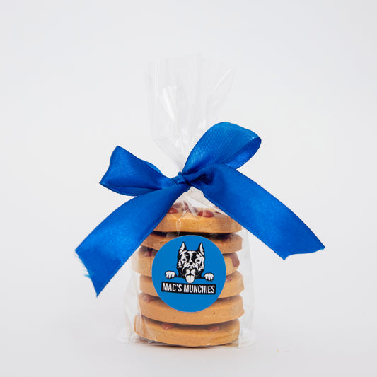 A stack of Cookie Bag treats from Macs Munchies, wrapped in clear plastic and tied with a large blue ribbon, features a round blue label with a dogs face illustration and the text MACS MUNCHIES, emphasizing that these cookies are made with dog-safe carob chips.