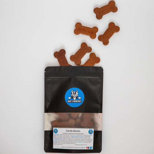 A package of Mac’s Munchies Carob Bones is open, revealing bone-shaped dog biscuits scattered on a white surface. The black packaging, featuring a blue logo with a dogs face, promotes gut health and is grain and wheat free.