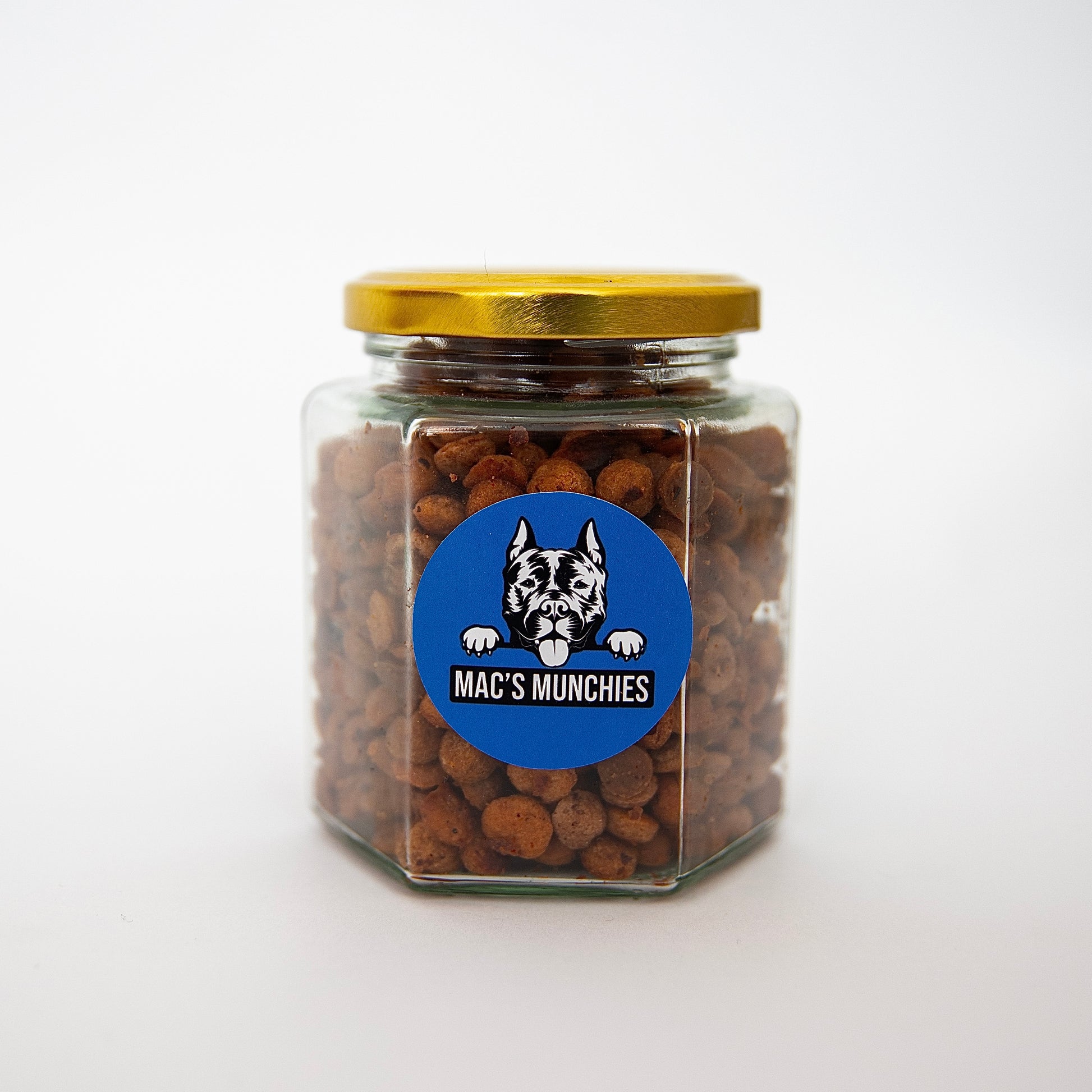 A hexagonal glass jar of Macs Munchies Training Treats, sealed with a gold lid, displays a blue sticker with a black and white dog illustration above the brand name. Made with low-fat peanut butter and banana, the jar sits on a plain white surface.