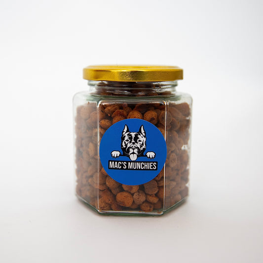 A hexagonal glass jar of Macs Munchies Training Treats, sealed with a gold lid, displays a blue sticker with a black and white dog illustration above the brand name. Made with low-fat peanut butter and banana, the jar sits on a plain white surface.