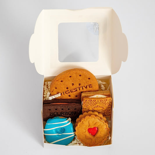 The Macs Munchies Classic British Biscuit Gift Box offers five cookies: a large digestive, a Bourbon, a round cookie with K9-safe blue icing, and two square cream-filled biscuits with patterns, all elegantly displayed against a crisp white background with an open lid.