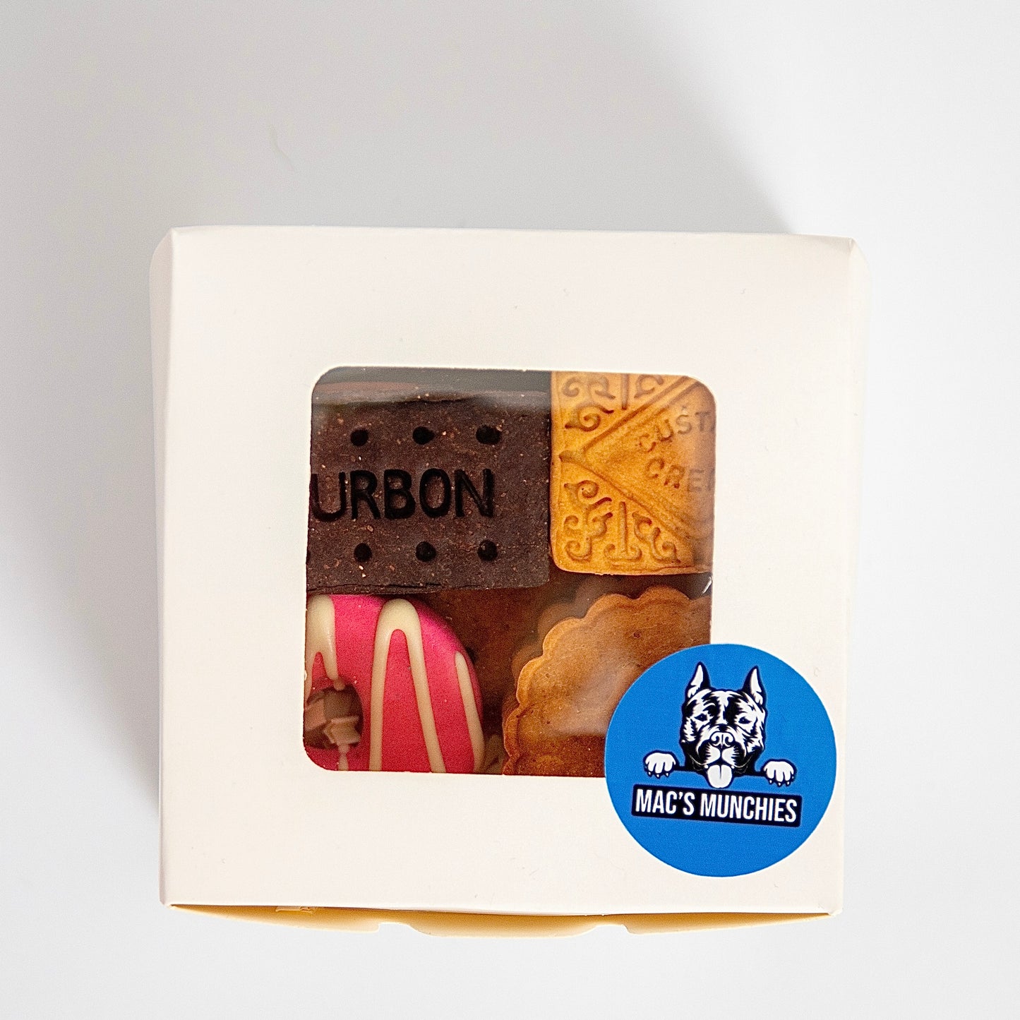 The Classic British Biscuit Gift Box from Macs Munchies features a clear window that showcases charming decorative dog treats, from bourbon biscuits to pink-striped delights, and boasts a blue circular label with a dogs face and MACS MUNCHIES, highlighting K9-safe icing.