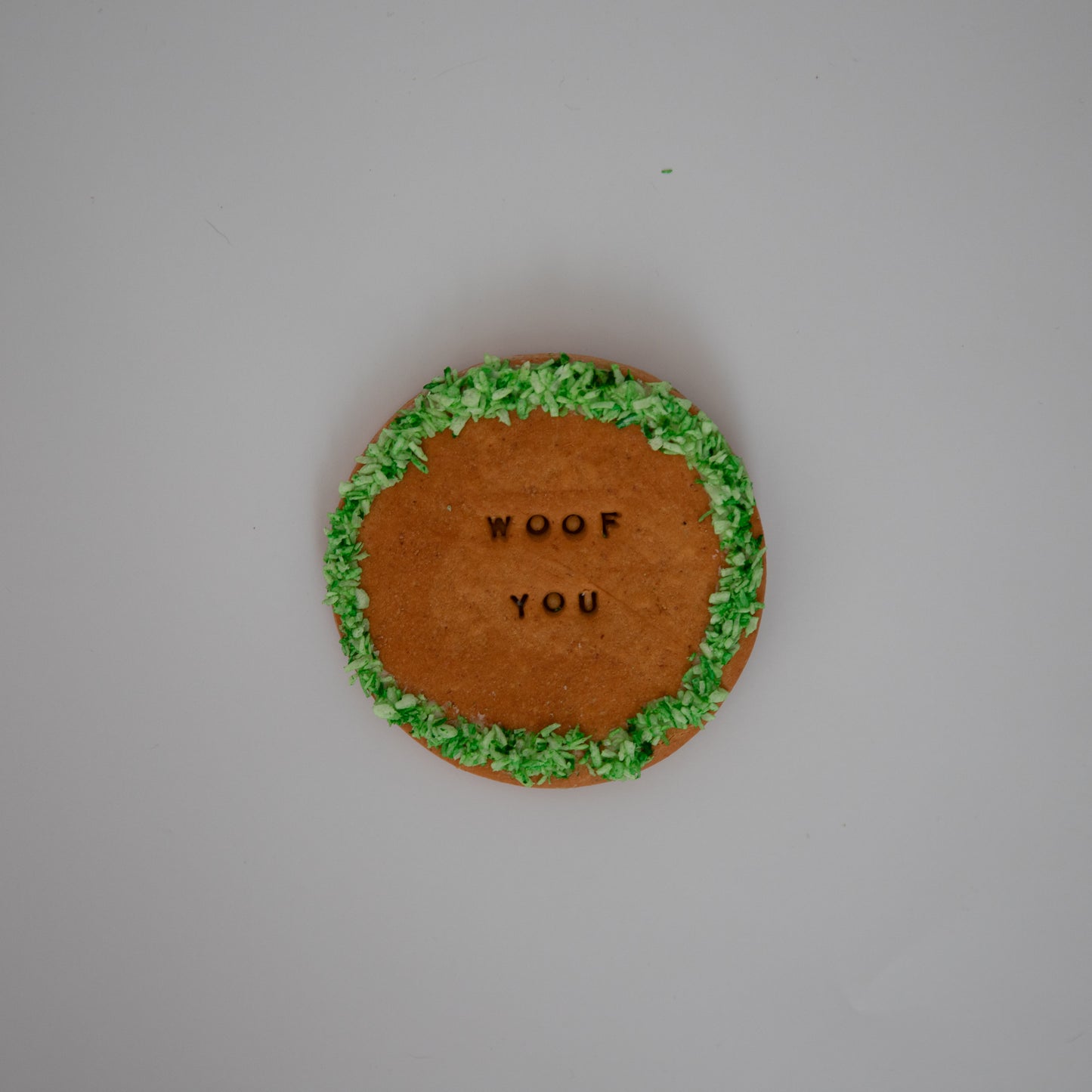 The XL Cookie by Macs Munchies is a round, peanut butter-flavored biscuit with WOOF YOU stamped on it and edged in green frosting, perfectly centered against a plain white background. Ideal as a hand-iced treat for your furry friend.