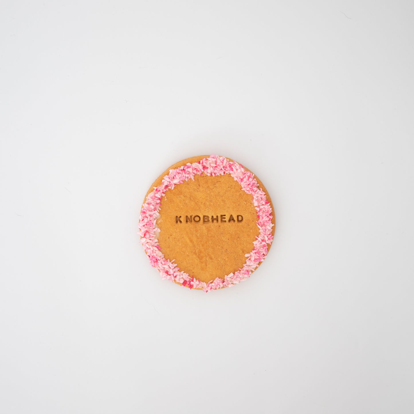 A round, XL peanut butter-flavored dog biscuit from Macs Munchies is adorned with pink icing on the edge, set against a white background. The word KNOBHEAD is crafted in the center of this hand-iced treat.