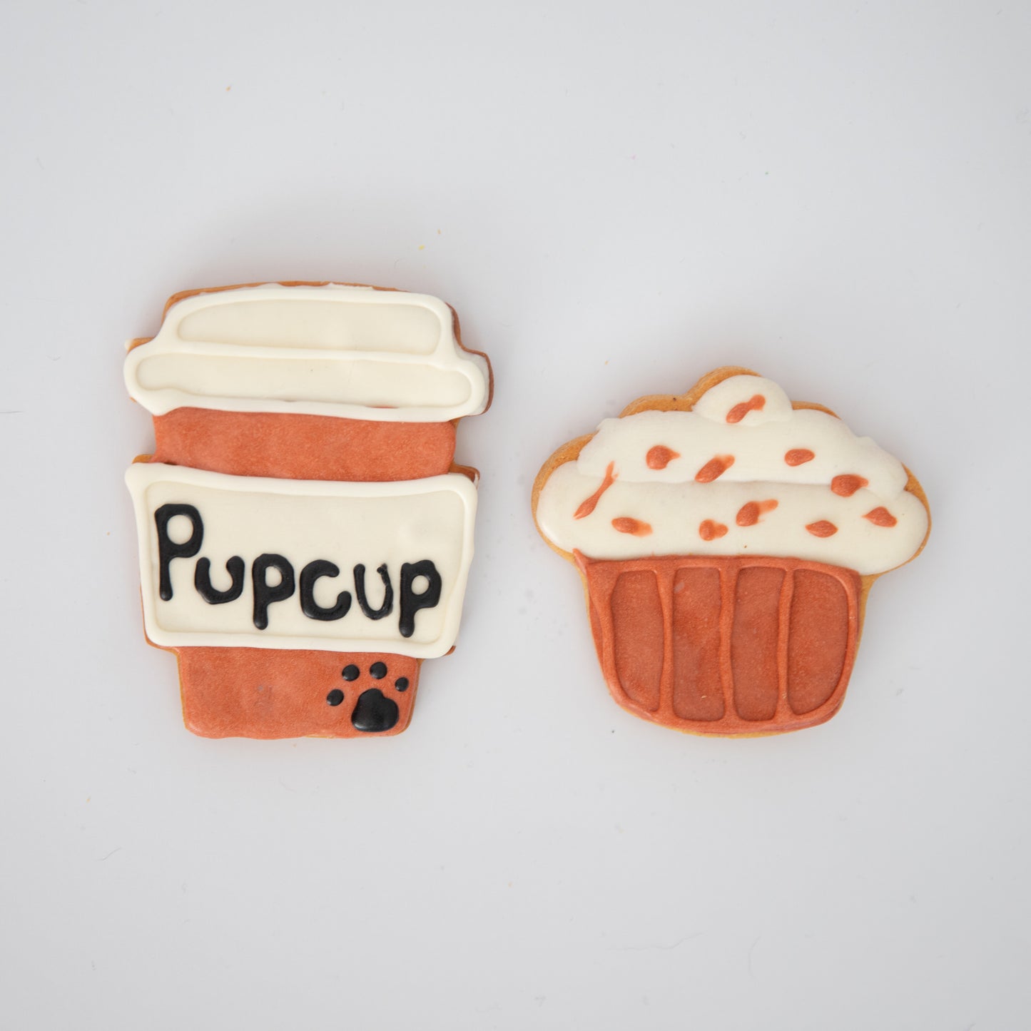 Macs Munchies Pupcup Pack includes two dog biscuits: a to-go coffee cup-shaped cookie with Pupcup and a paw print, and a cupcake-shaped one with K9-safe icing and orange sprinkles, both offering a delicious peanut butter flavor.