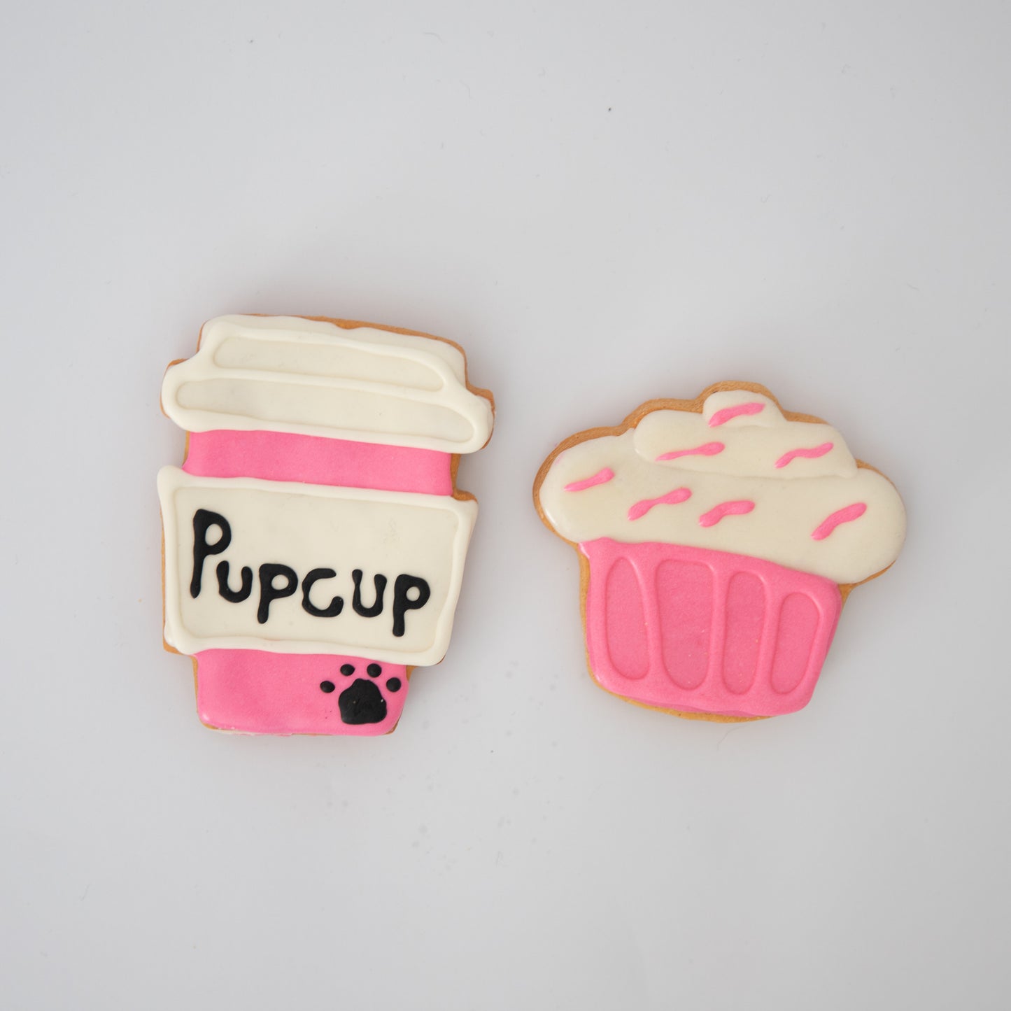 Macs Munchies Pupcup Pack features two delightful cookies: a Pupcup coffee cup with a pink sleeve and black paw print, and a cupcake with K9-safe icing, pink sprinkles, and peanut butter flavor – perfect treats for canine companions!.