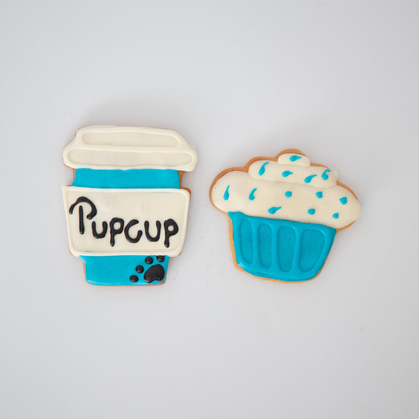 The Pupcup Pack by Macs Munchies features two peanut butter-flavored dog treats: one is a blue coffee cup with Pupcup label, K9-safe icing, and a white lid; the other is a blue cupcake with white icing and sprinkles, set against a plain white background.